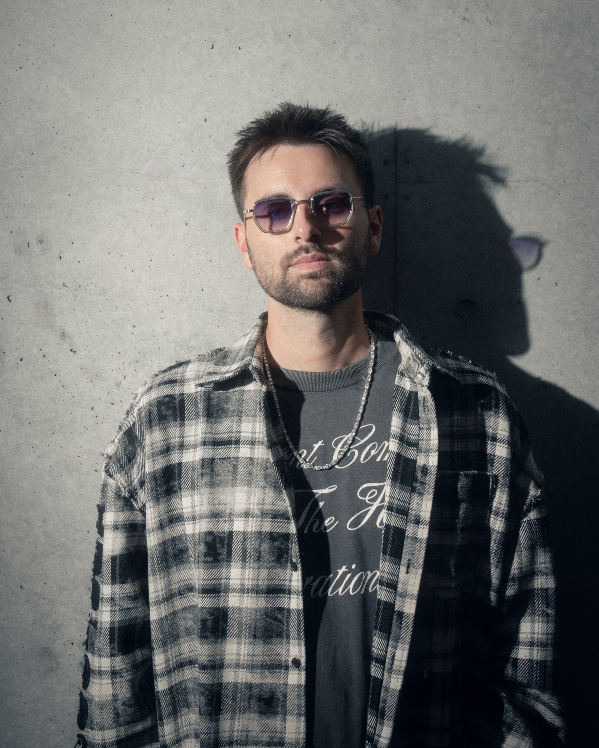 GATANO against a concrete wall in hard sunlight, in a checked overshirt and tinted glasses.